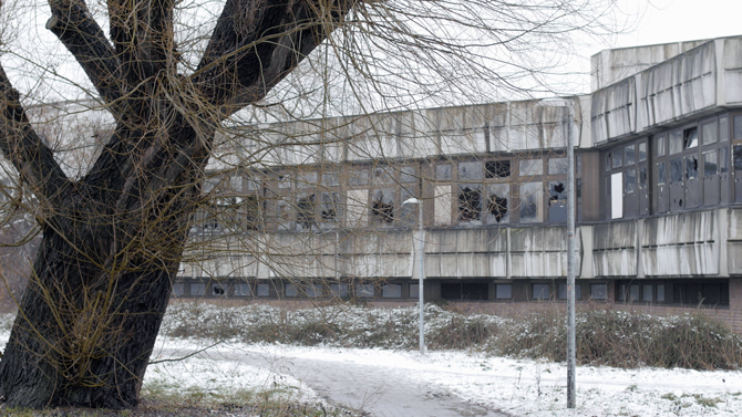 Das Bild zeigt ein verlassenes Gebäude aus dreckigem Sichtbeton mit zerschlagenen Fensterfronten. Vor dem Gebäude steht ein Baum ohne Blätter. Ein Weg vor dem Gebäude und Büssche die von etwas Schnee bedeckt sind.